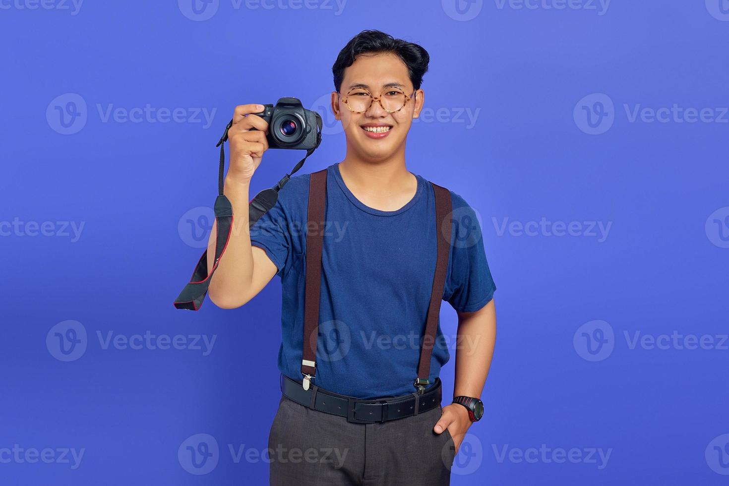 beau photographe montrant un appareil photo professionnel et souriant joyeusement sur fond violet