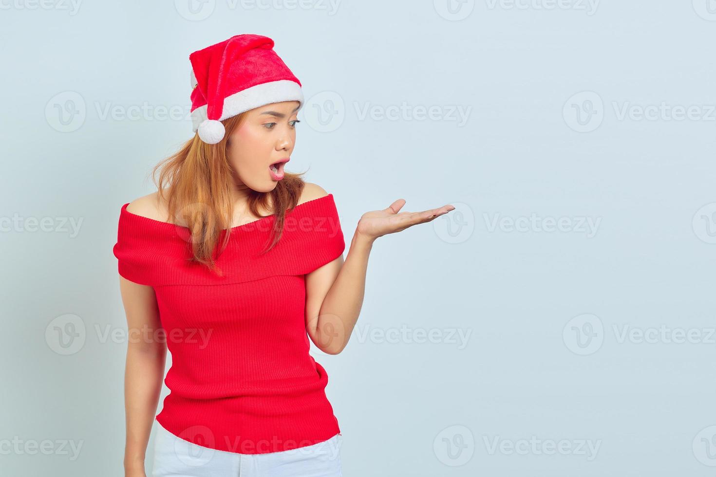 Portrait de jeune femme asiatique surprise au chapeau de Noël et montrant l'espace de copie dans la paume sur fond blanc photo