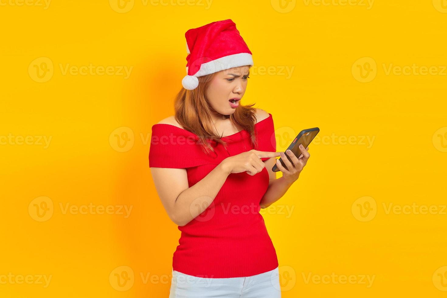 surpris, jeune, femme asiatique, dans, santa claus, chapeau, doigt pointé, à, téléphone portable, isolé, sur, fond jaune photo