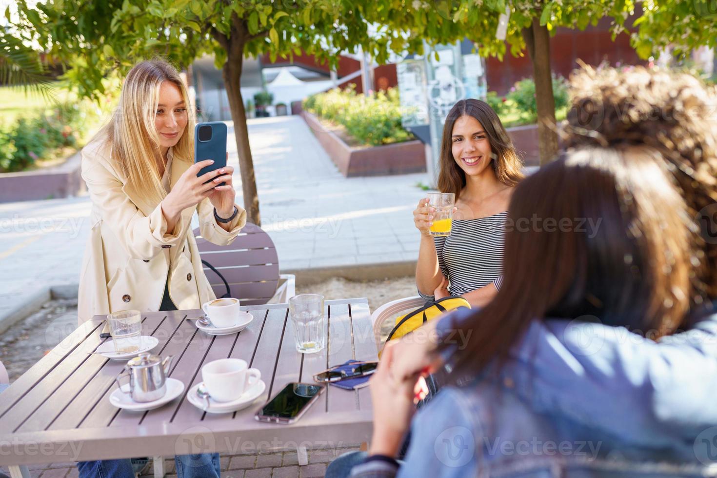 jeune fille blonde prenant une photo avec un smartphone de ses amis assis à une table de bar.