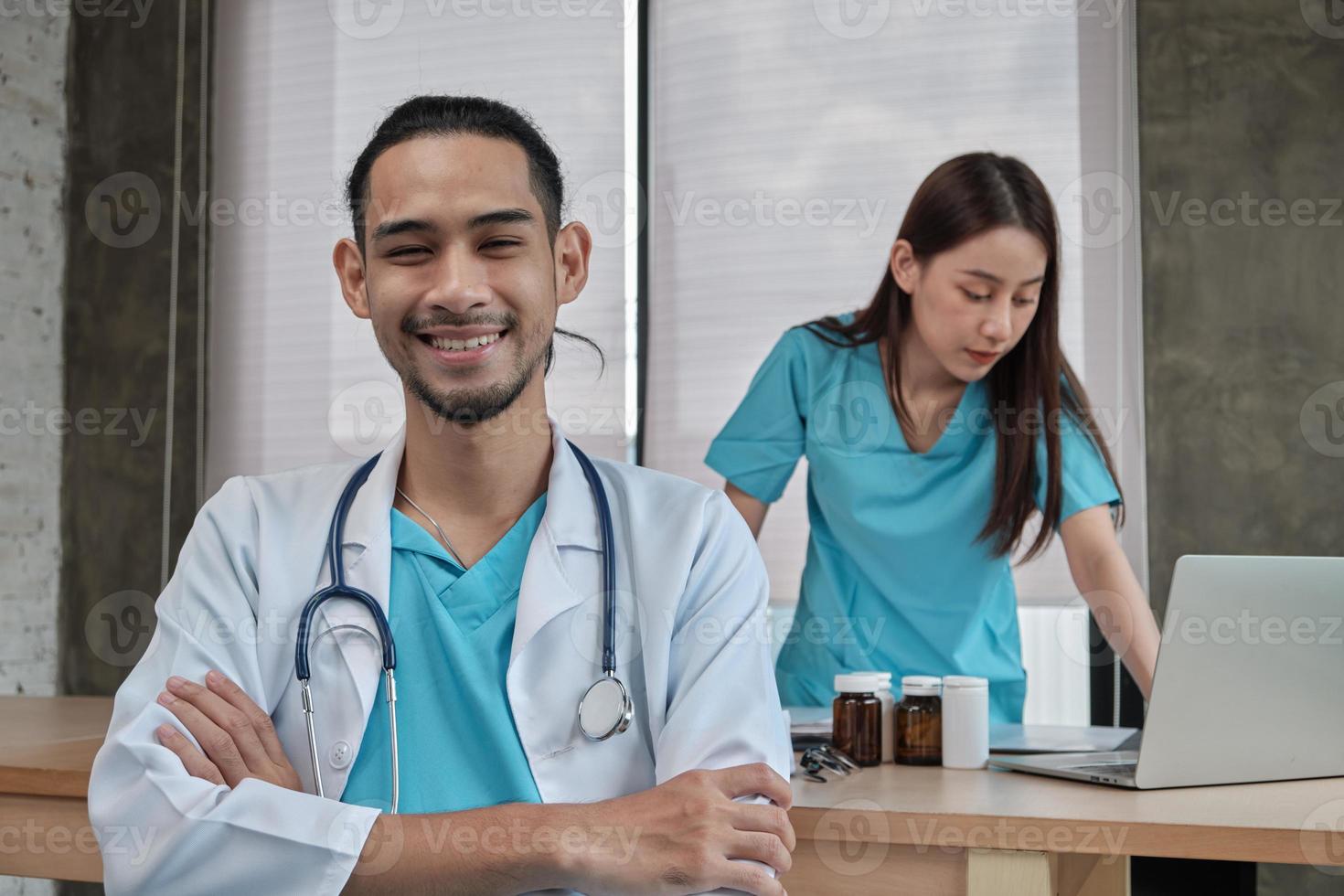 portrait d'un jeune médecin d'origine asiatique en uniforme avec stéthoscope. sourire et regarder la caméra dans une clinique hospitalière, partenaire féminin travaillant derrière, professionnel de la médication à deux personnes. photo