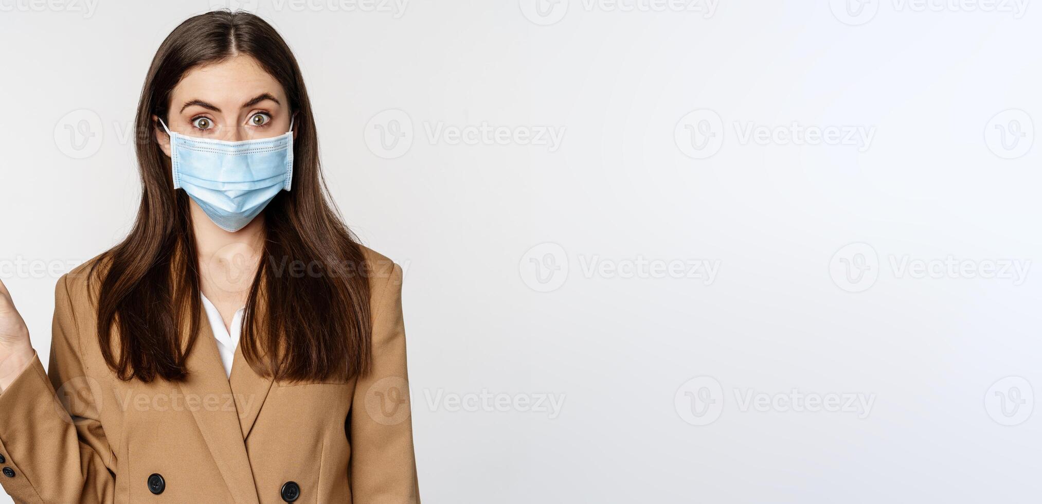 coronavirus et gens concept. portrait de affaires femme à lieu de travail portant visage masque, montrer du doigt doigt la gauche à logo, entreprise bannière, blanc Contexte photo