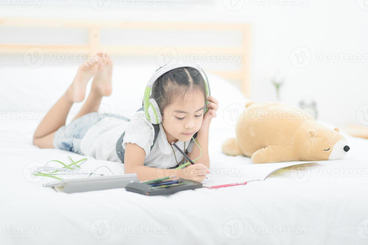 jolie petite fille asiatique faisant ses devoirs, écrivant avec des crayons colorés sur le lit à la maison. design élégant pour les enfants qui jouent, l'apprentissage préscolaire et le concept d'éducation artistique créative. photo