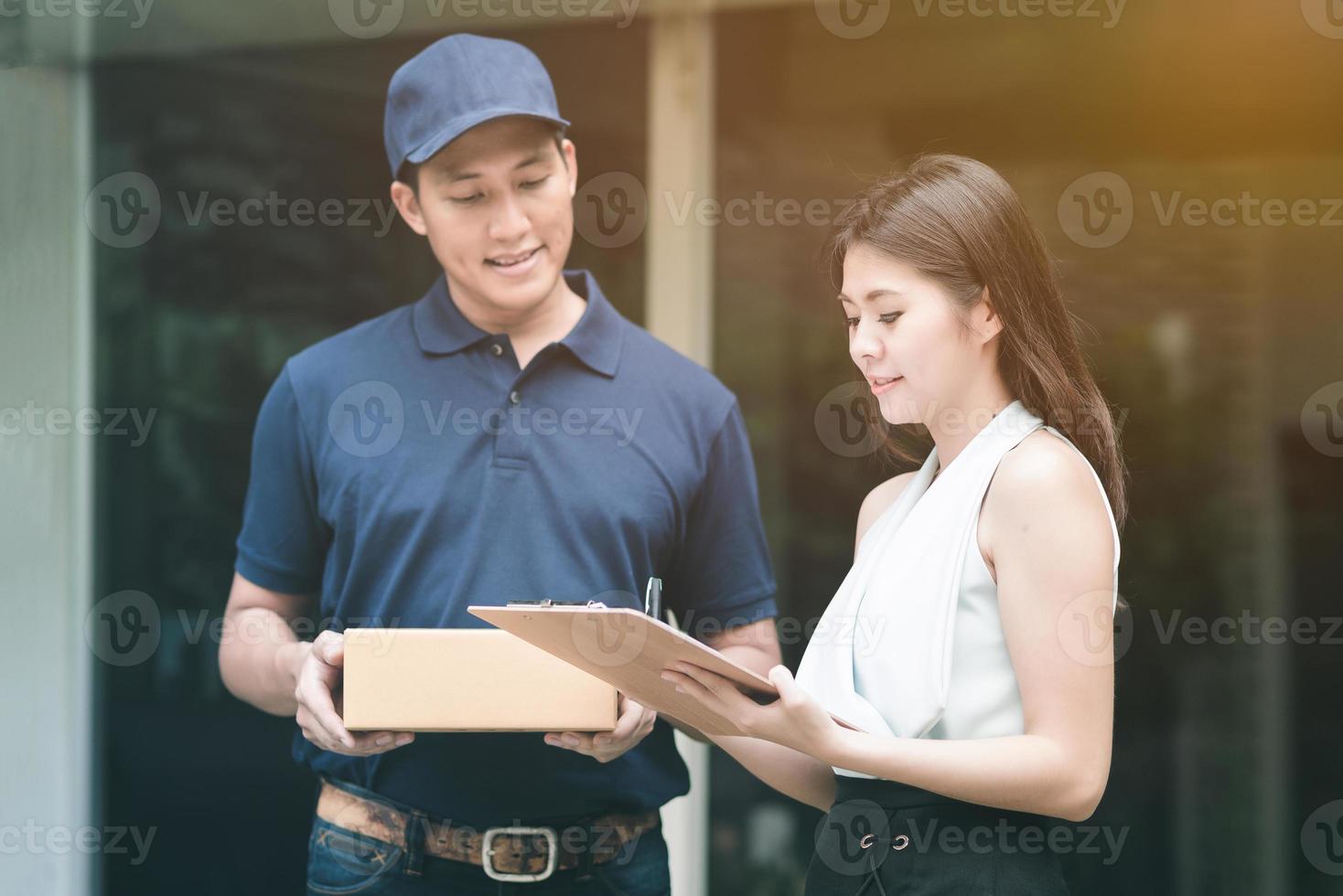 beau jeune livreur asiatique souriant et tenant une boîte en carton en attendant la belle jeune femme asiatique mettant la signature dans la tablette intelligente pour confirmer la réception. photo