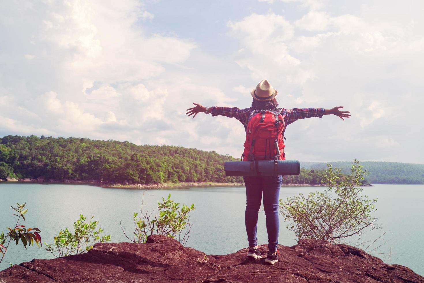 jeune femme hipster debout sur la pierre au bord du lac montre la liberté des mains et profite de la nature des voyageurs photo
