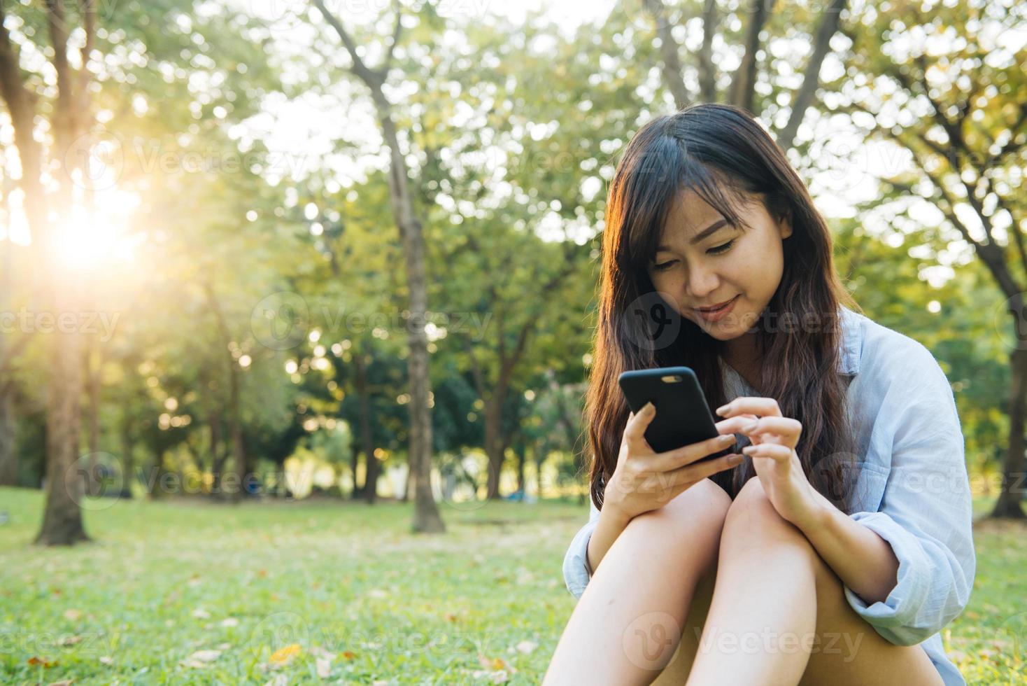 jolie femme asiatique lisant un message texte agréable sur son téléphone portable alors qu'elle était assise dans le parc le jour du printemps. femme asiatique utilisant un téléphone intelligent avec un sentiment de détente et un visage souriant. concepts de style de vie et de technologie. photo