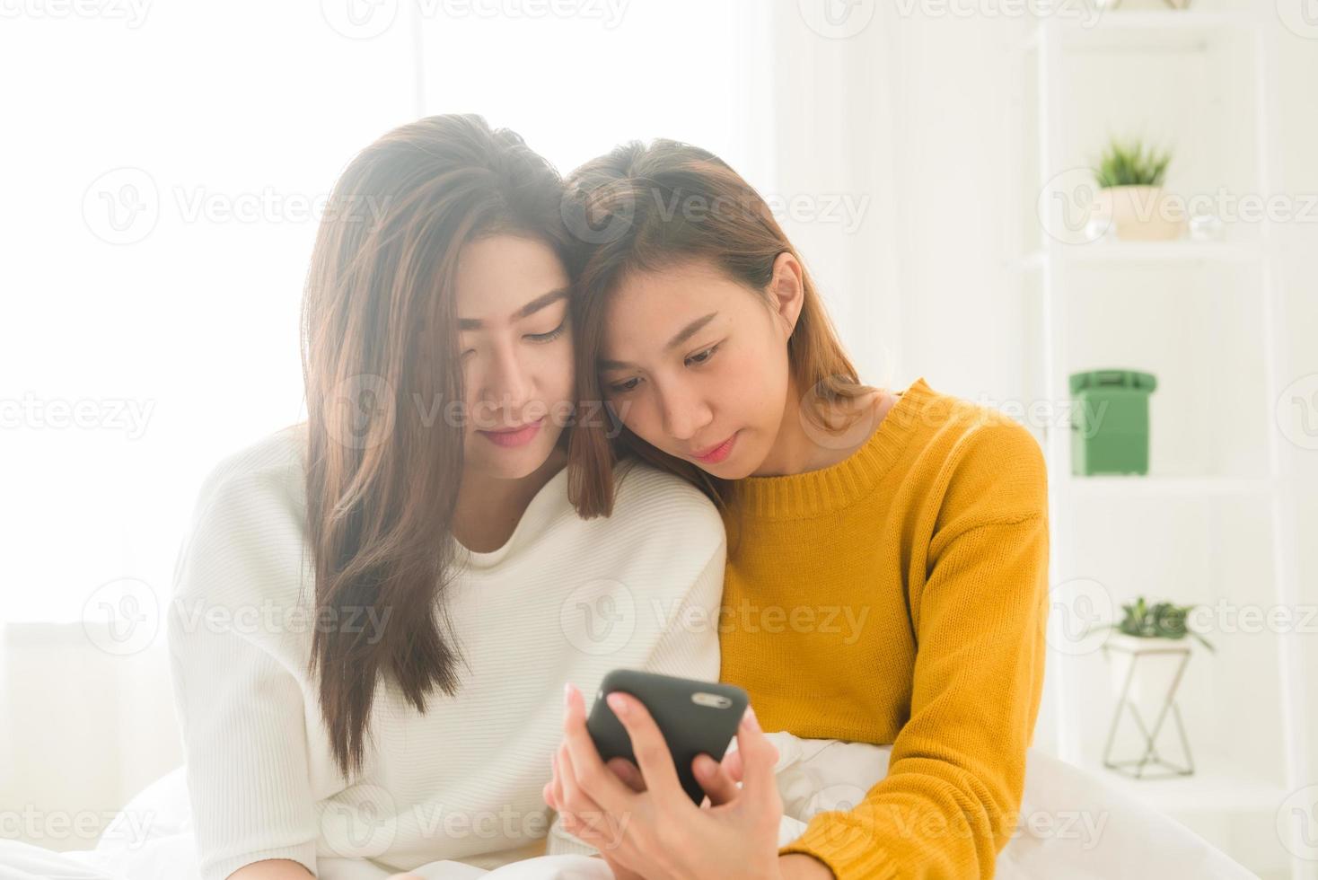 belles jeunes femmes asiatiques lgbt couple heureux lesbien assis sur le lit étreinte et utilisant le téléphone ensemble chambre à coucher à la maison. couple de lesbiennes lgbt ensemble à l'intérieur concept. passer du bon temps à la maison. photo