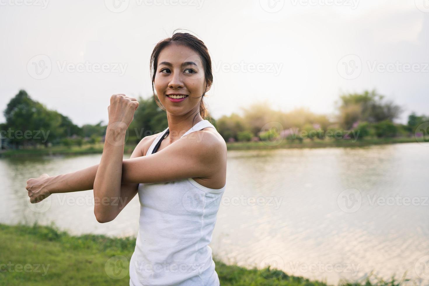 Une jeune femme asiatique en bonne santé réchauffe le corps en s'étirant avant l'exercice et le yoga près du lac au parc sous une lumière chaude et légère le matin. mode de vie fitness et femmes actives s'exercent dans le concept de ville urbaine. photo