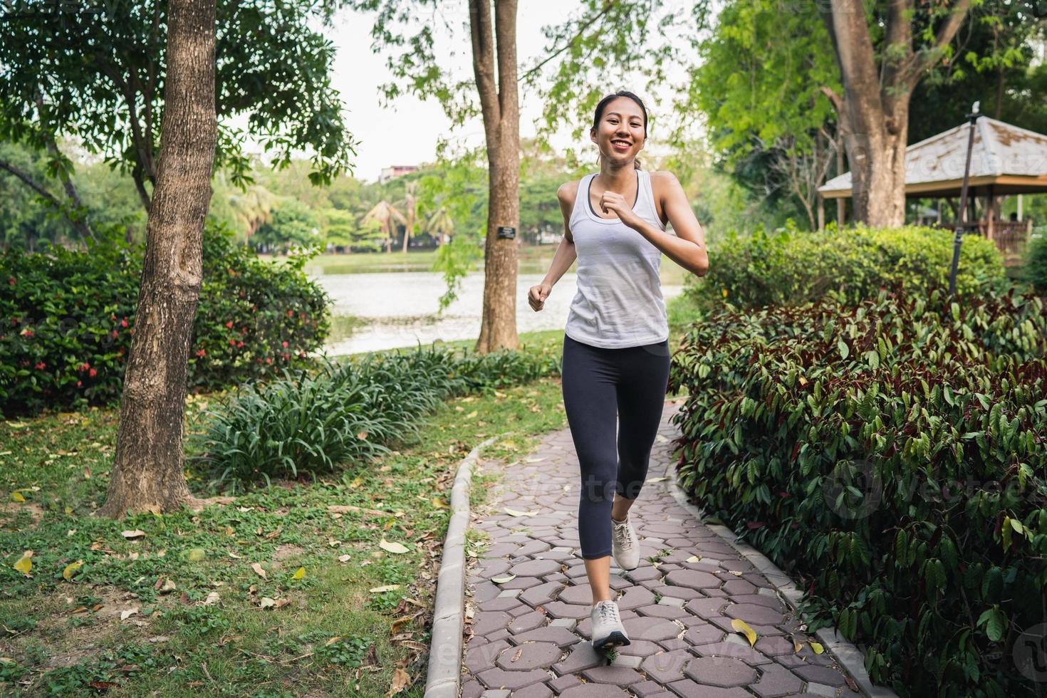 belle jeune femme asiatique en bonne santé en vêtements de sport courir et faire du jogging sur le trottoir près du lac au parc le matin. mode de vie fitness et femmes actives s'exercent dans le concept de ville urbaine. photo