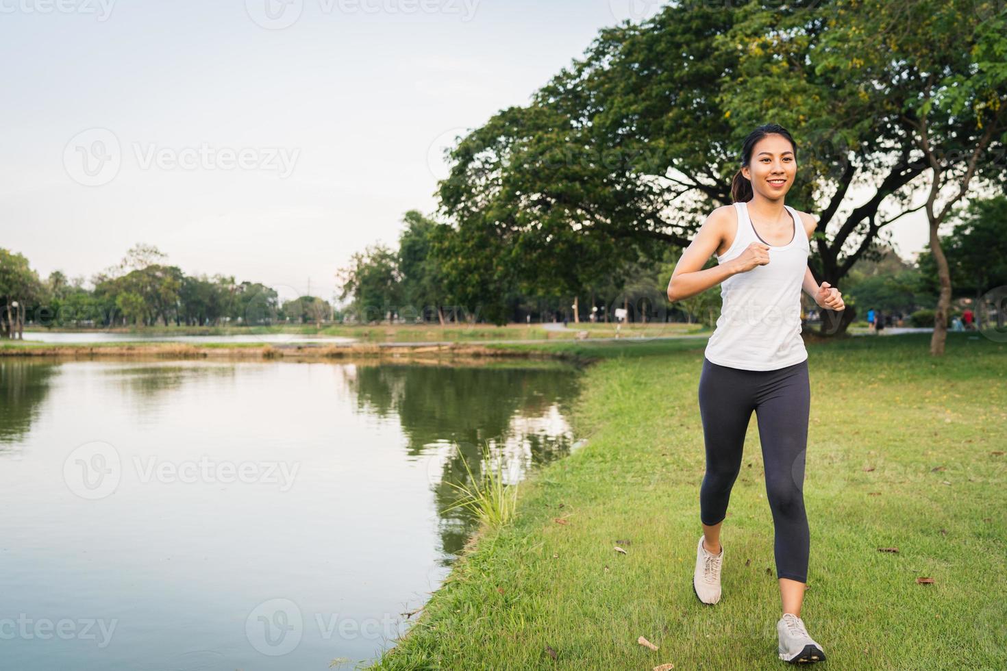 belle jeune femme asiatique en bonne santé en vêtements de sport courir et faire du jogging sur le trottoir près du lac au parc le matin. mode de vie fitness et femmes actives s'exercent dans le concept de ville urbaine. photo