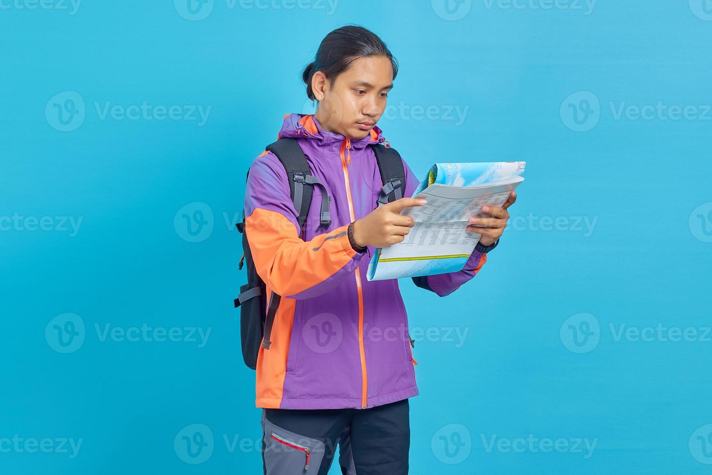 Portrait d'un jeune homme asiatique portant une veste et un sac de transport à la lecture d'une carte de lecture sérieuse isolée sur fond bleu photo