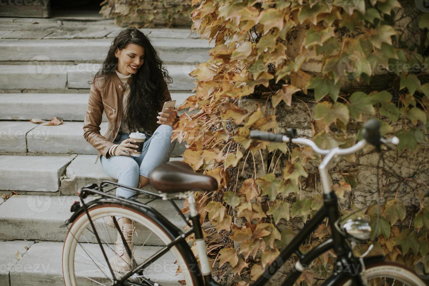 jeune femme avec téléphone portable boire du café à emporter dans les escaliers par le vélo le jour de l'automne photo