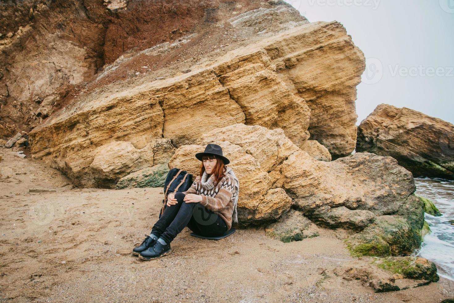 femme dans un chapeau et une écharpe avec un sac à dos est assise sur la côte sur fond de rochers contre une mer magnifique photo