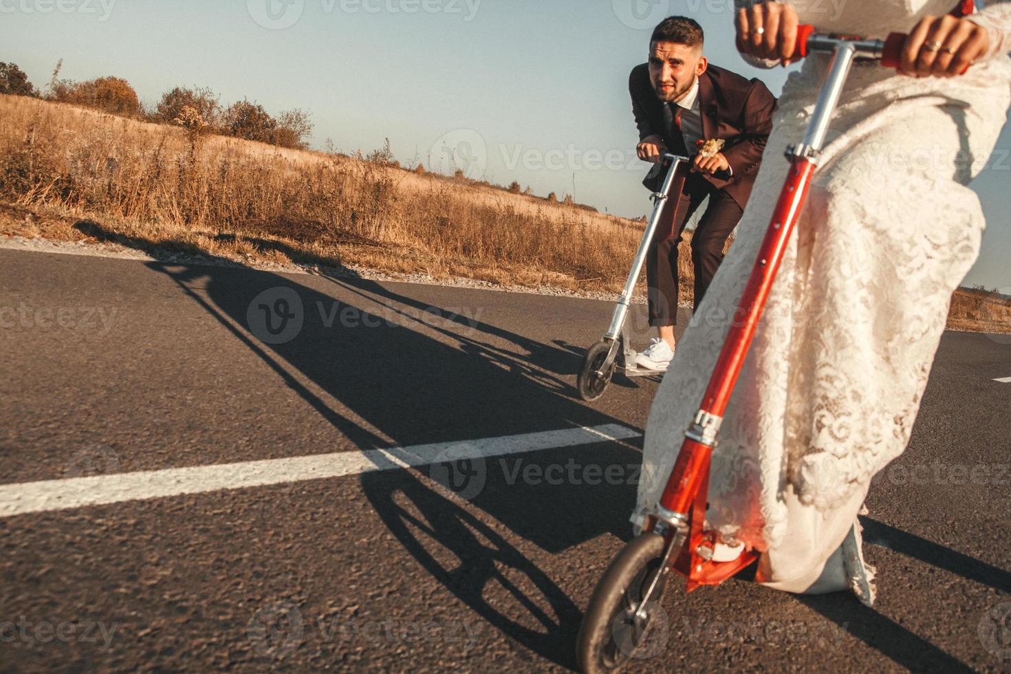 couple de mariage souriant chevauchant un scooter le long de la route à l'extérieur de la ville au coucher du soleil. place pour le texte ou la publicité photo