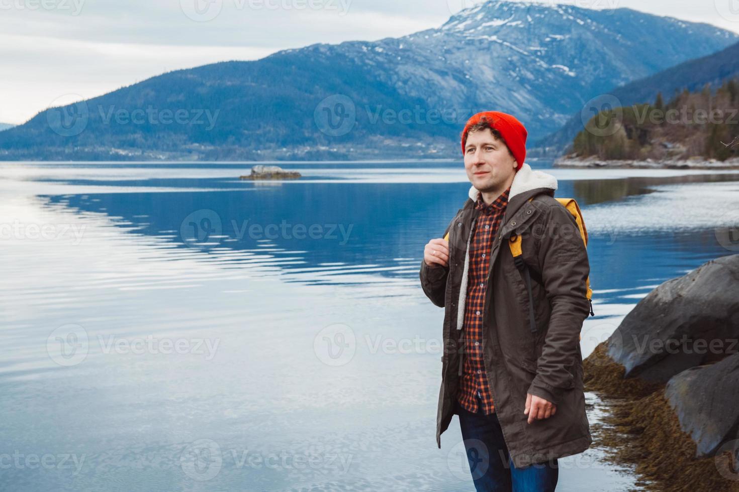 jeune homme avec un sac à dos jaune portant un chapeau rouge debout sur un rocher sur fond de montagne et de lac. espace pour votre message texte ou contenu promotionnel. concept de mode de vie de voyage photo