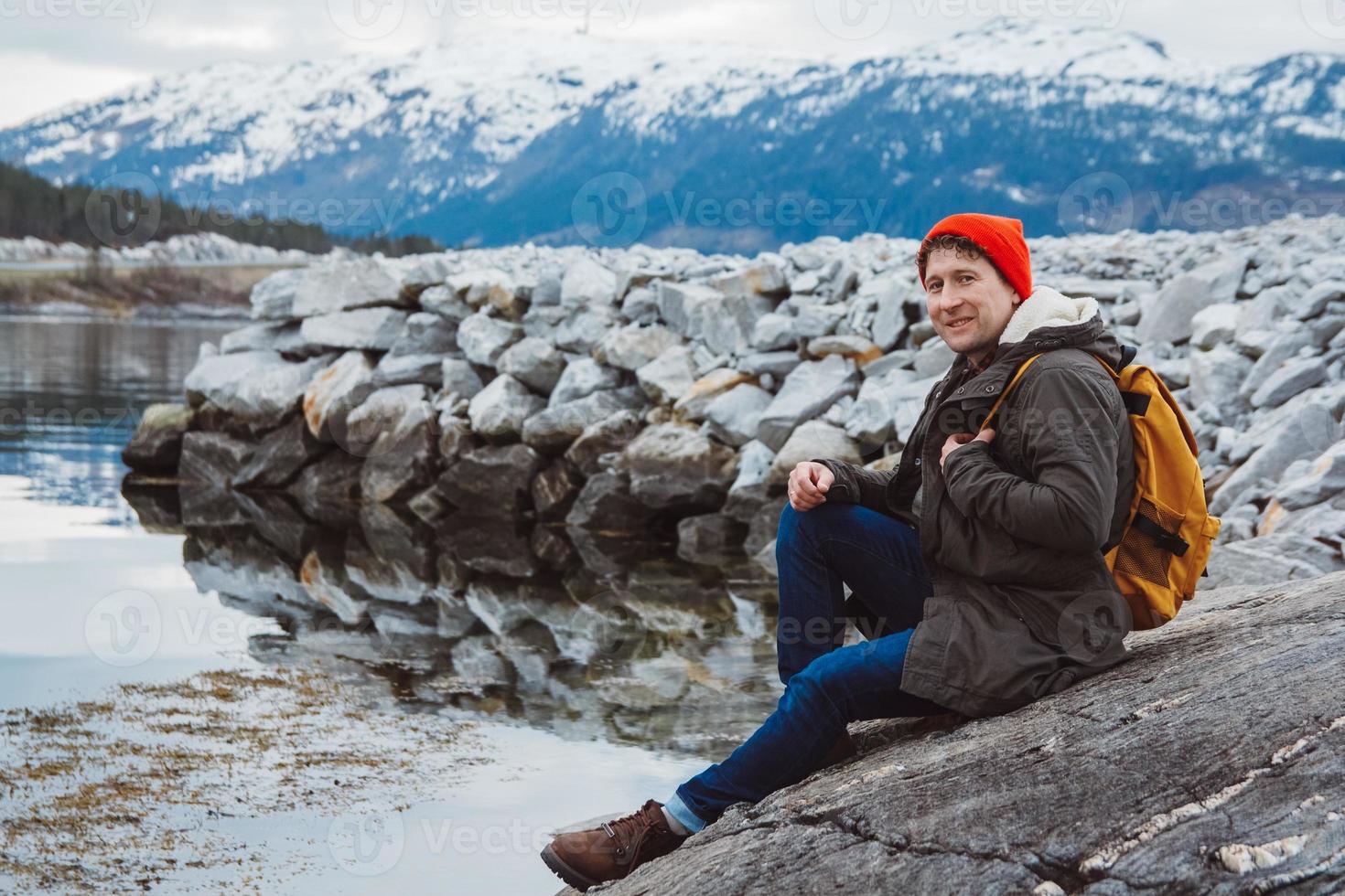 jeune homme avec un sac à dos jaune portant un chapeau rouge assis sur le rivage sur fond de montagne et de lac. espace pour votre message texte ou contenu promotionnel. concept de mode de vie de voyage. photo