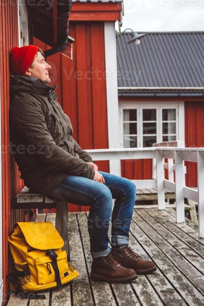 homme voyageur avec un sac à dos jaune portant est assis près de la maison en bois de couleur rouge. concept de mode de vie de voyage. photo