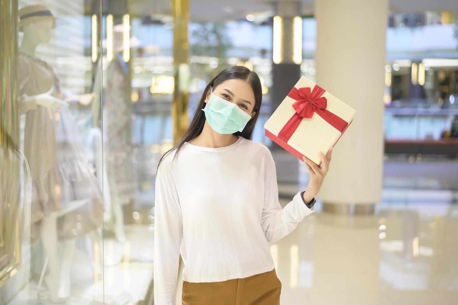 femme portant un masque de protection tenant une boîte-cadeau dans un centre commercial, faisant du shopping sous le concept de pandémie de covid-19, d'action de grâces et de noël. photo