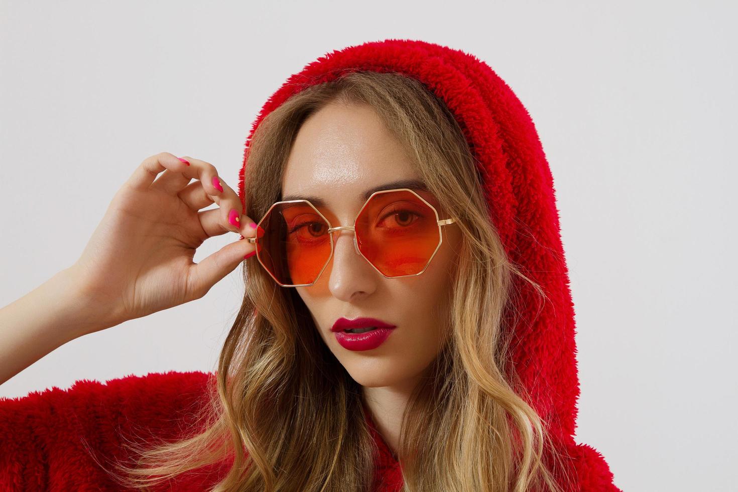 visage de femme gros plan dans des lunettes de soleil rouges isolés sur fond blanc. fille en sweat à capuche rouge et maquillage des lèvres rouges. concept de beauté de mode. belle peau de visage et concept d'accessoires à la mode. photo