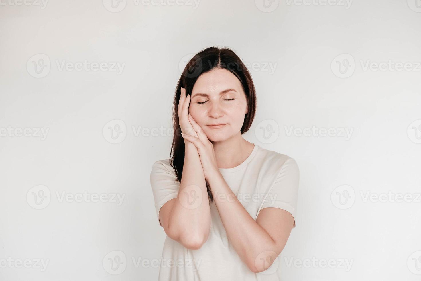 une femme aux yeux fermés garde les mains sur la joue, vêtue d'un t-shirt blanc décontracté, a un maquillage minimal, pose à l'intérieur sur un fond blanc photo