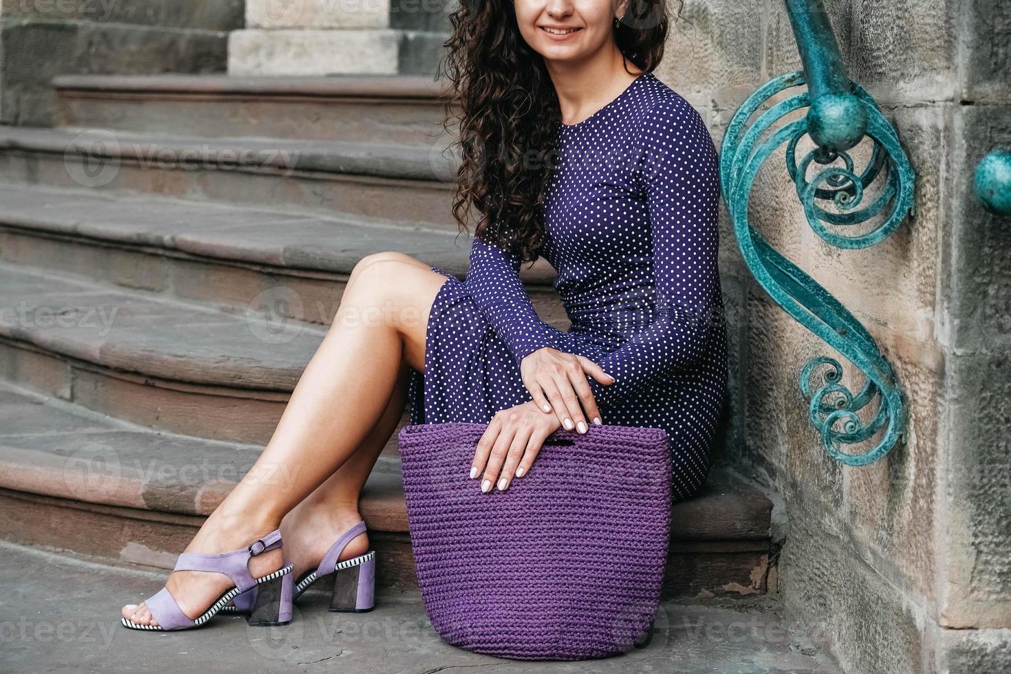 femme portant une robe violette à pois, tenant un sac tricoté assis sur les escaliers posant dans la rue de la ville. copie, espace vide pour le texte photo