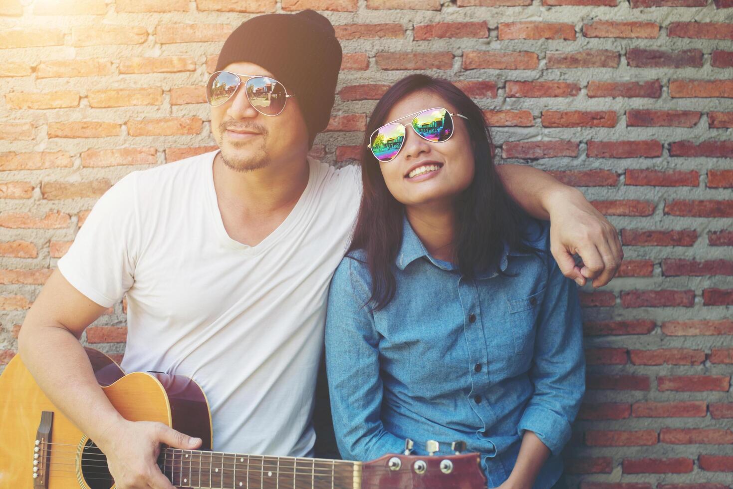 un couple hipster se serre dans ses bras, regarde au loin et sourit tout en se tenant debout en plein air jouant de la guitare contre un mur de briques, les rencontres ont passé du bon temps ensemble. photo