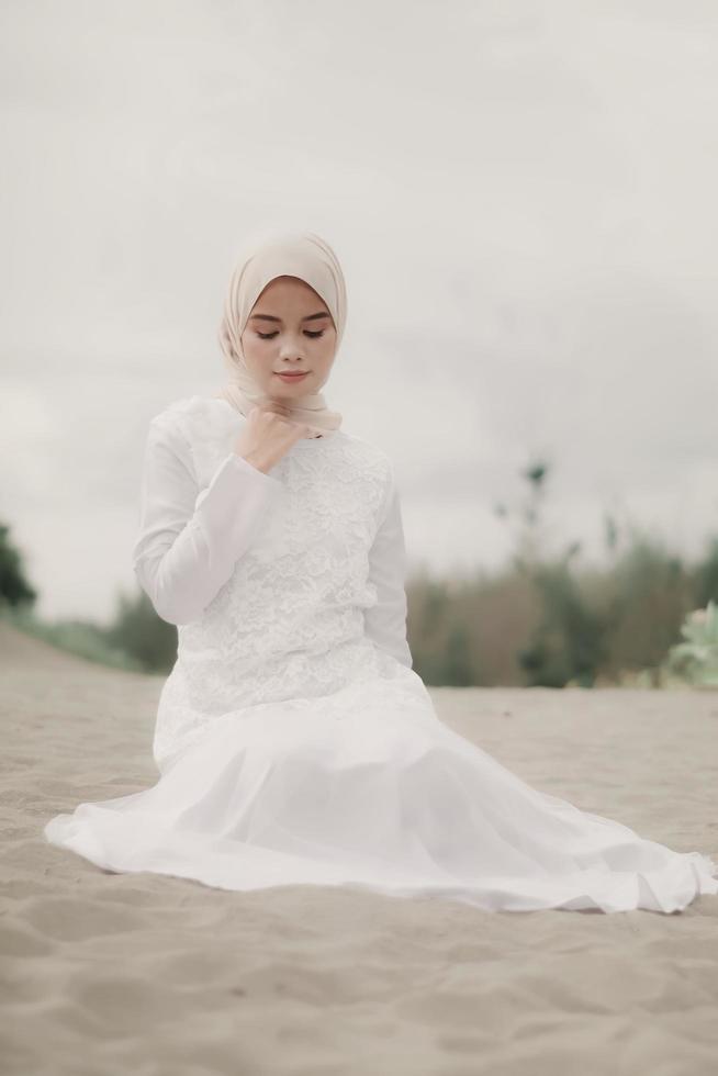 beau modèle féminin islamique portant la mode hijab, une robe de mariée moderne pour femme musulmane assise dans le sable et la plage. portrait d'une fille asiatique modèle utilisant le hijab s'amusant à la plage avec des arbres photo