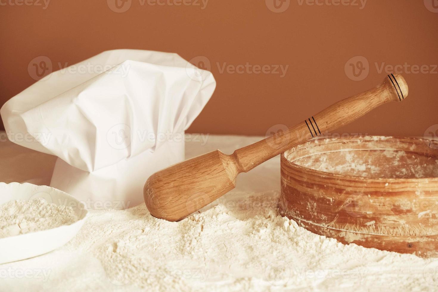 farine de blé blanche éparpillée sur une table, rouleau à pâtisserie en bois, tamis rustique rond et chapeau de cuisinier sur fond marron. cuisiner de délicieux biscuits dans la cuisine photo