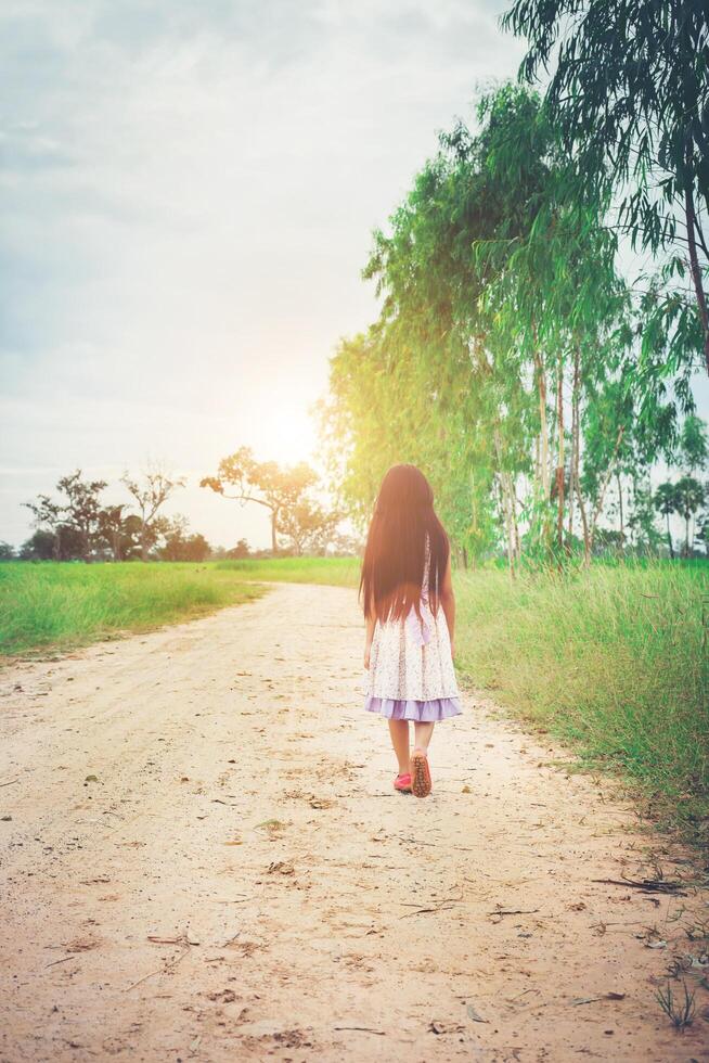 une petite fille aux cheveux longs en robe s'éloigne de vous sur une route rurale. photo