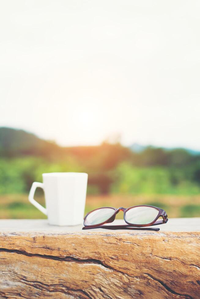 blanc de tasse de café chaud avec des verres sur le banc avec fond de montagne nature verdoyante. photo