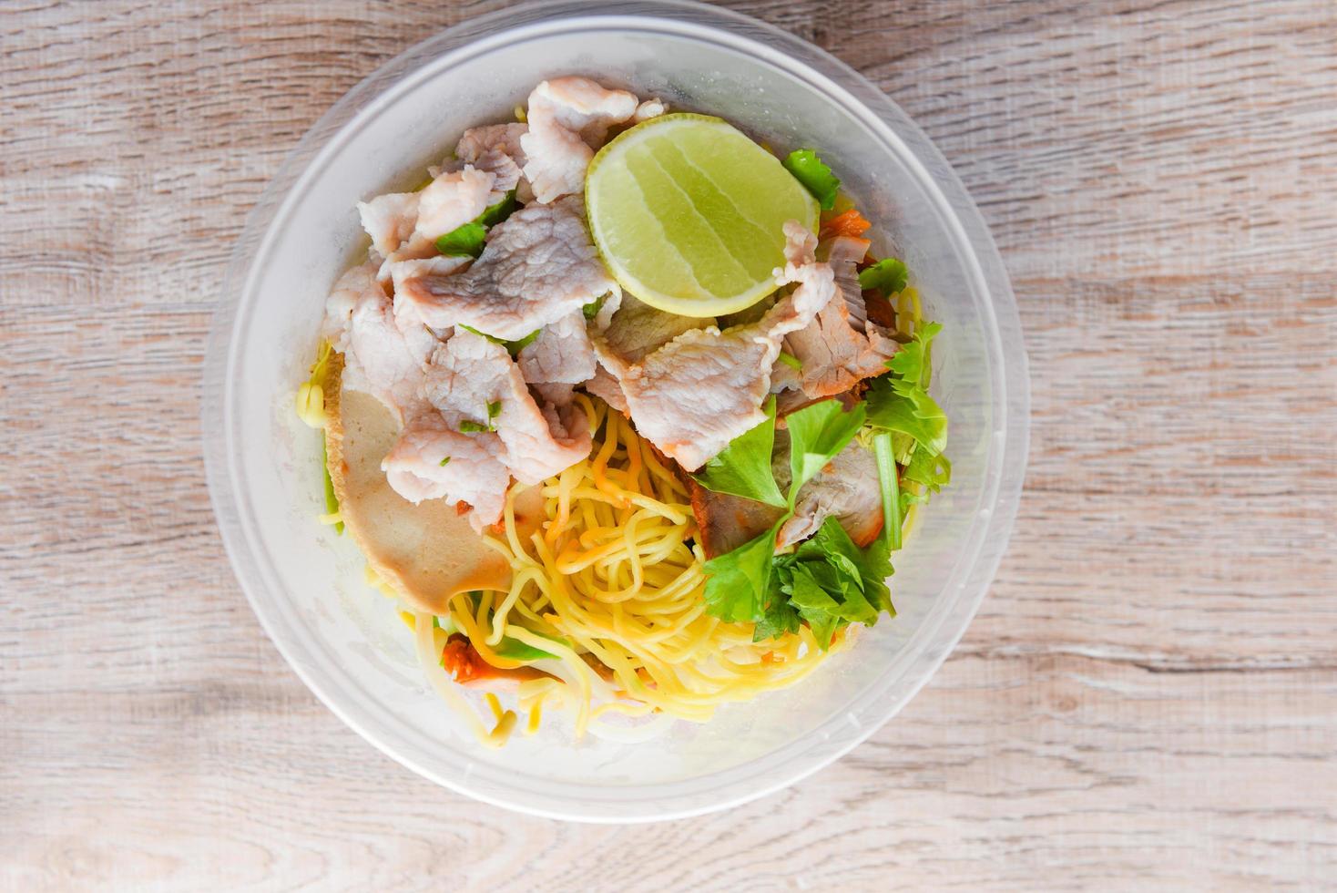 vue de dessus thaï et chine cuisine asiatique nouilles jaunes citron citron vert, nouilles sèches porc avec porc rôti rouge et légumes dans un bol en plastique sur une table en bois photo