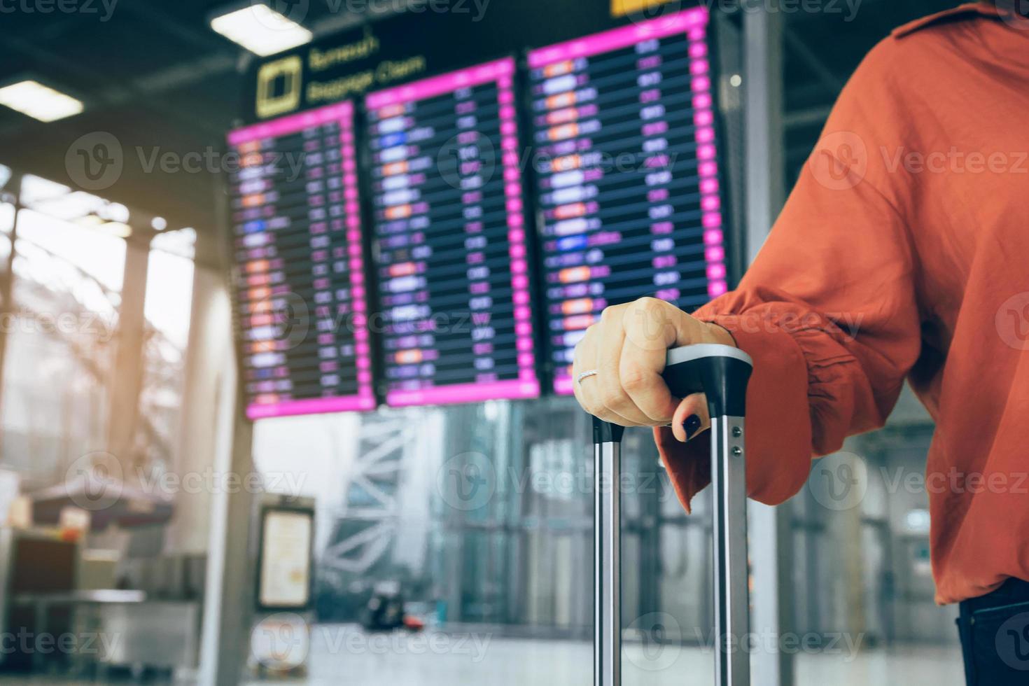 jeune femme asiatique debout à l'aéroport tenant une valise vérifiant le départ avec l'horaire de vol en arrière-plan. photo