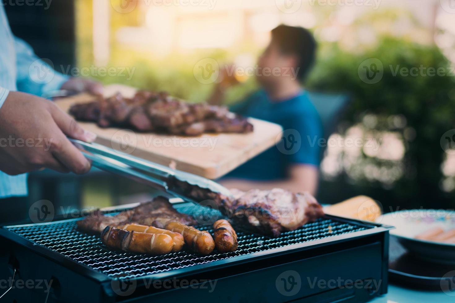 gros plan homme main asiatique pince le porc sur le gril et le tient à des amis qui célèbrent dans le dos. photo