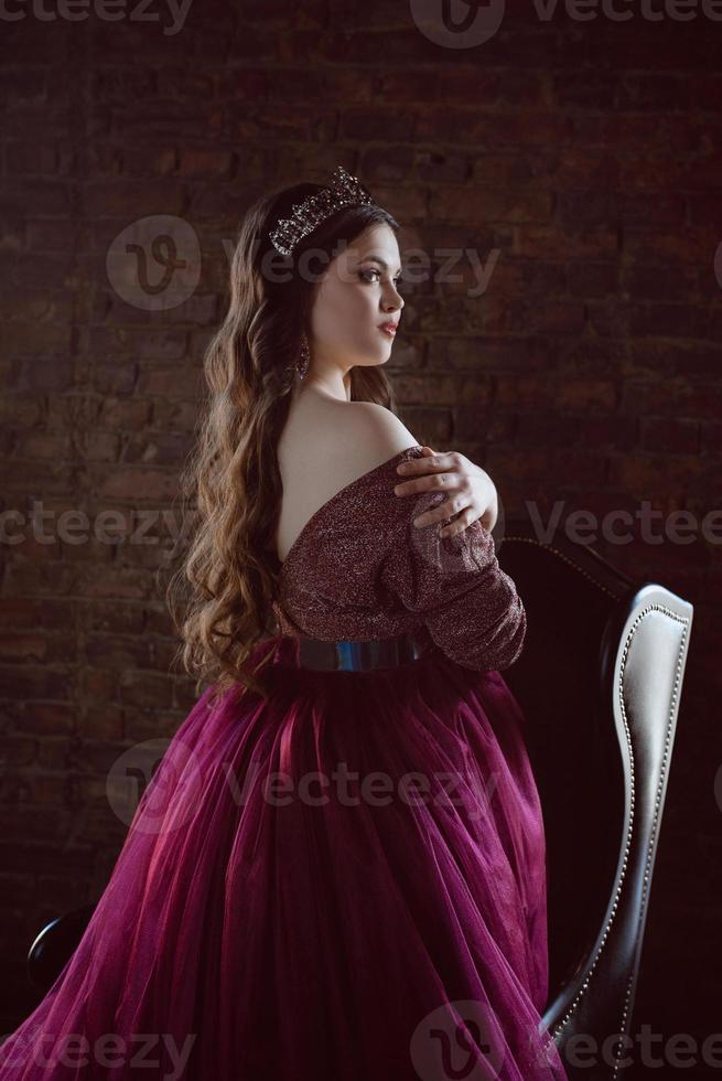 portrait de belle jeune princesse jolie jolie femme en longue robe et couronne de reine violette, avec de longs cheveux et maquillage intérieur à l'intérieur du loft près d'une chaise en cuir photo