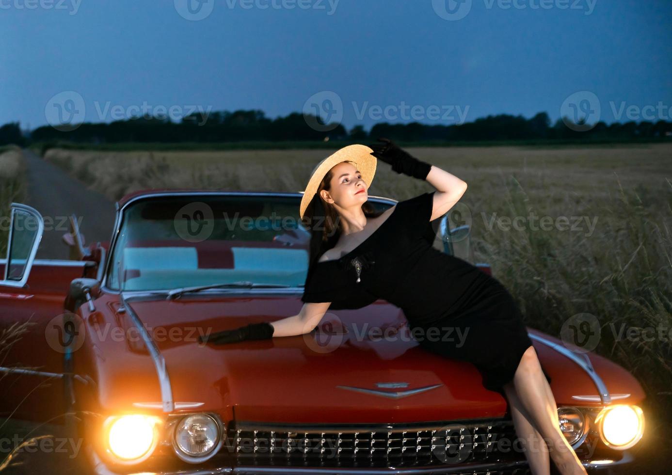 une belle fille pose sur le capot d'un caddy convertible rouge, soir, coucher de soleil. photo