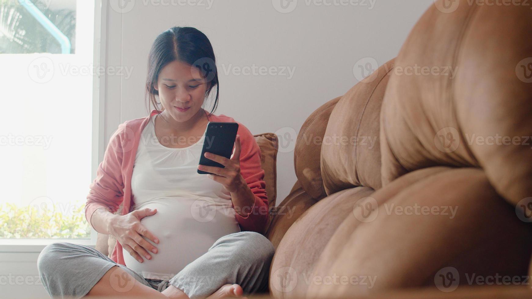 jeune femme enceinte asiatique utilisant un téléphone portable pour rechercher des informations sur la grossesse. maman se sentant heureuse souriante positive et paisible tout en prenant soin de son enfant allongé sur un canapé dans le salon à la maison concept. photo