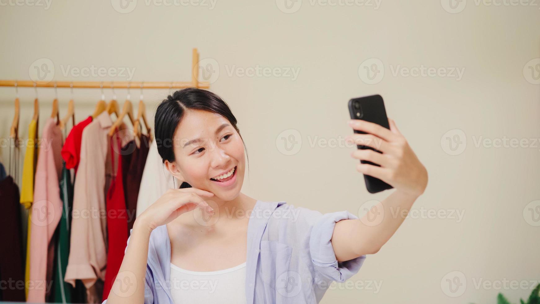 heureuse belle jeune femme asiatique à l'aide de smartphone selfie en direct en ligne sur le réseau social. beauté blogueuse présente des cosmétiques de beauté assis devant la caméra de la tablette pour enregistrer une vidéo. photo