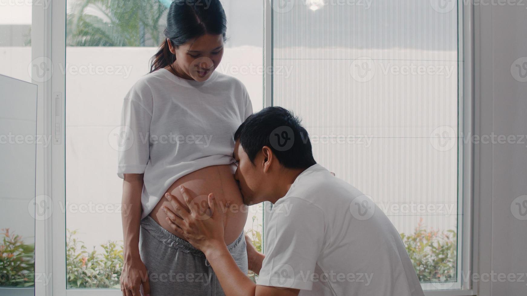 jeune couple enceinte asiatique homme embrassant le ventre de sa femme parlant avec son enfant. maman et papa se sentent heureux souriant paisiblement tout en prenant soin de bébé, grossesse près de la fenêtre dans le salon à la maison concept. photo