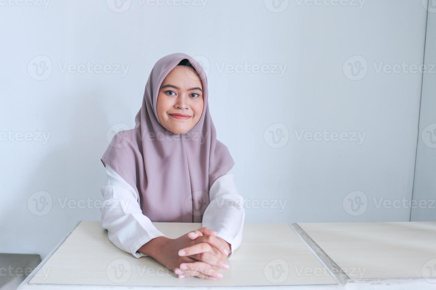 une jeune femme islamique asiatique portant un foulard est assise sur la table en se tenant la main et en souriant. islam femme indonésienne sur fond gris photo