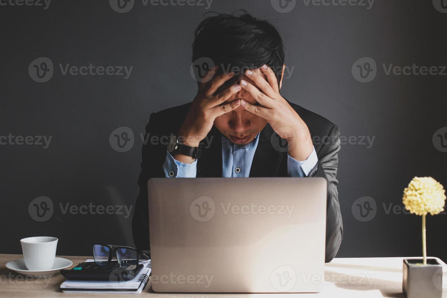 homme d'affaires asiatique stressé tenant la tête tout en cherchant des idées avec un ordinateur portable, une tasse à café, une calculatrice sur le livre, des plantes décoratives et un livre sur un bureau en bois isolé sur fond gris photo