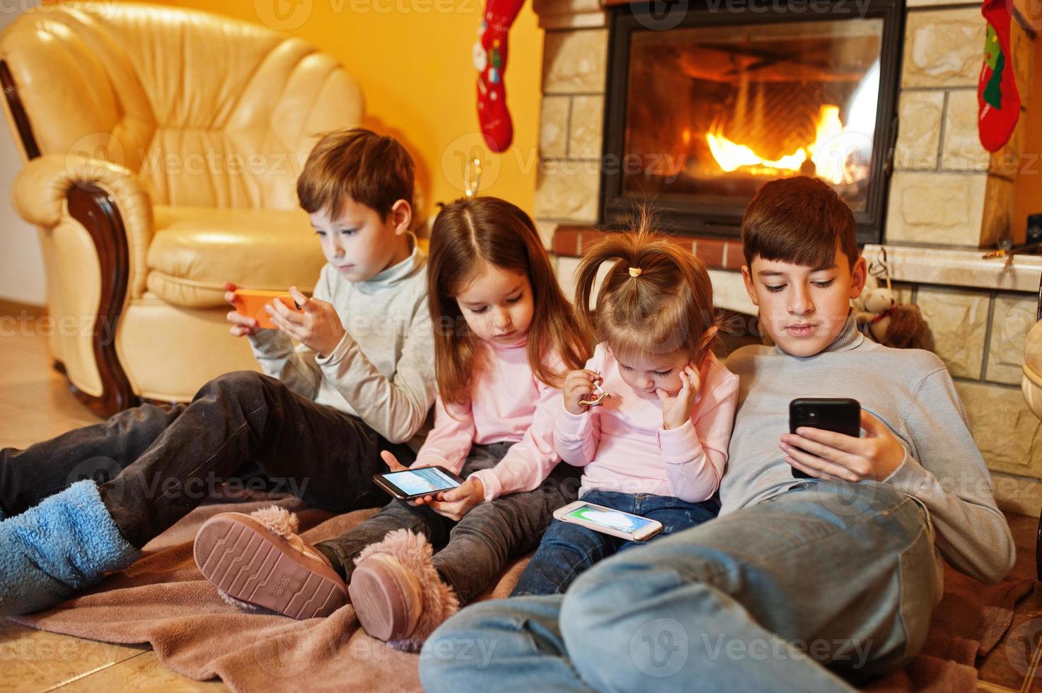 heureux quatre enfants qui regardent dans leur téléphone portable à la maison près d'une cheminée dans un salon chaleureux le jour de l'hiver. photo