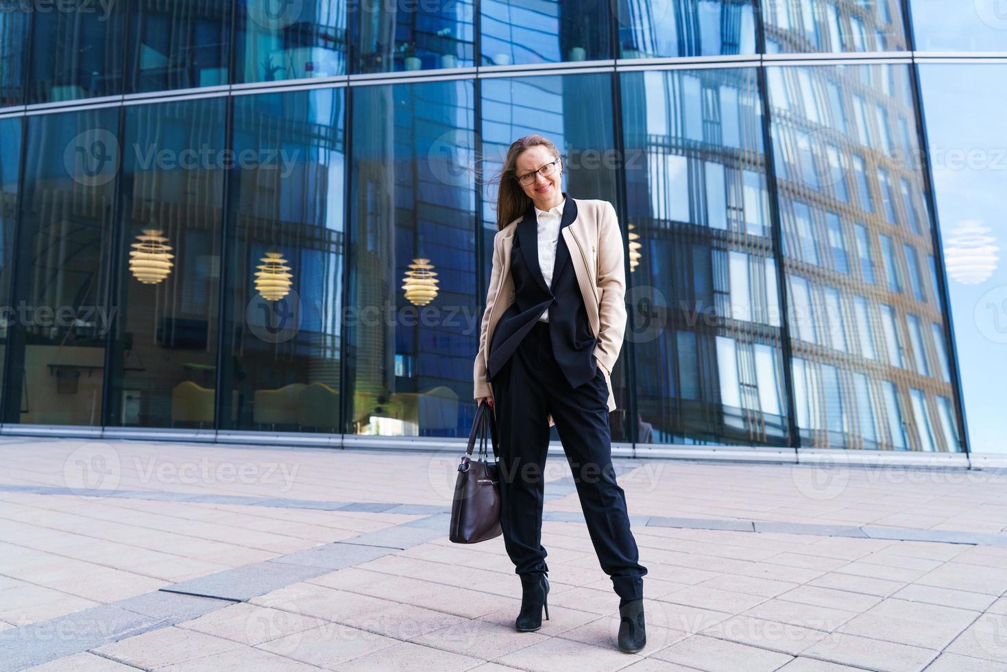 femme réussie avec des lunettes se tient devant un immeuble de bureaux avec sac photo