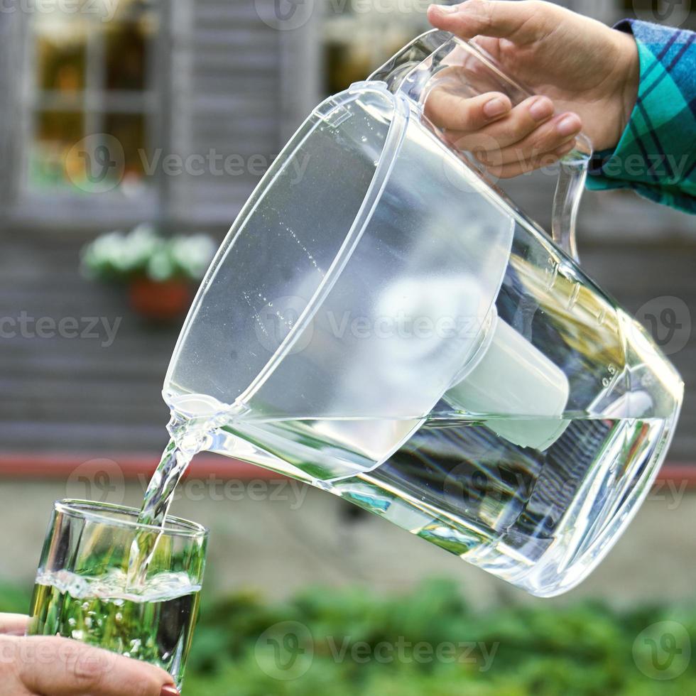 la main d'une fille verse de l'eau filtrée claire d'une cruche de filtration d'eau dans un verre que la main féminine tient devant une maison de campagne photo