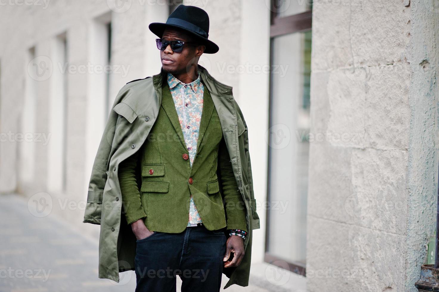 portrait de mode d'un homme afro-américain noir sur une veste en velours vert, un chapeau noir et un manteau sur ses épaules marchant dans les rues de la maison de fond de la ville avec de nombreuses fenêtres photo