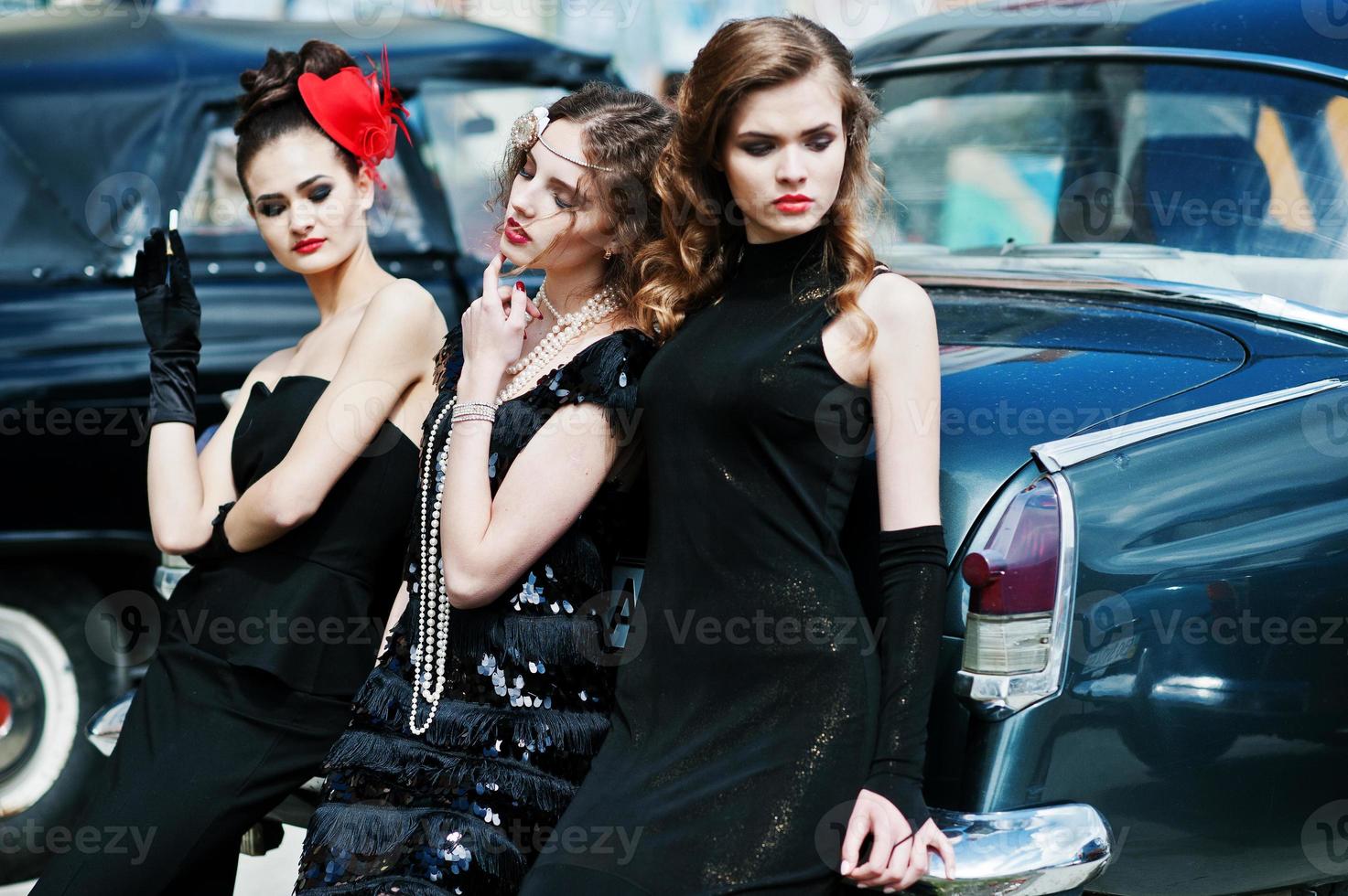 trois jeunes filles en robe de style rétro près de vieilles voitures vintage classiques. photo