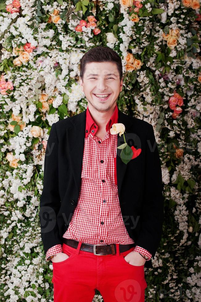 mode jeune homme riant avec rose, en costume noir et pantalon rouge pose décontractée au studio, mur de décor floral de fond. photo