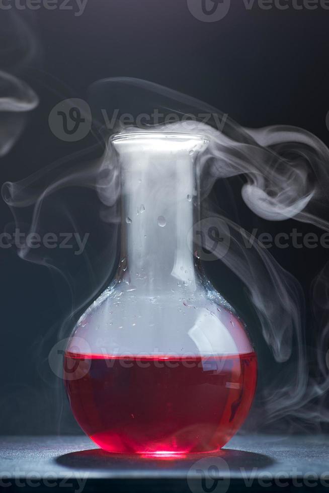 une photo verticale d'une fiole en verre avec un liquide rouge fume sur fond bleu.