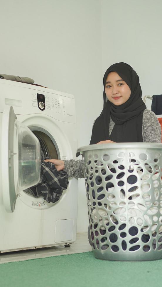 une femme asiatique en hijab met des vêtements sales dans un lave-linge à la maison photo