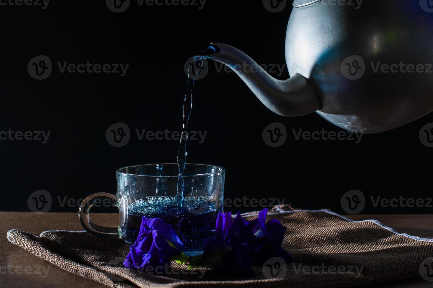 versez le thé aux pois papillons par un pot en métal dans une tasse avec une fleur violette sur une nappe marron sur fond noir. boisson saine à boire. herbes et concept médical. photo