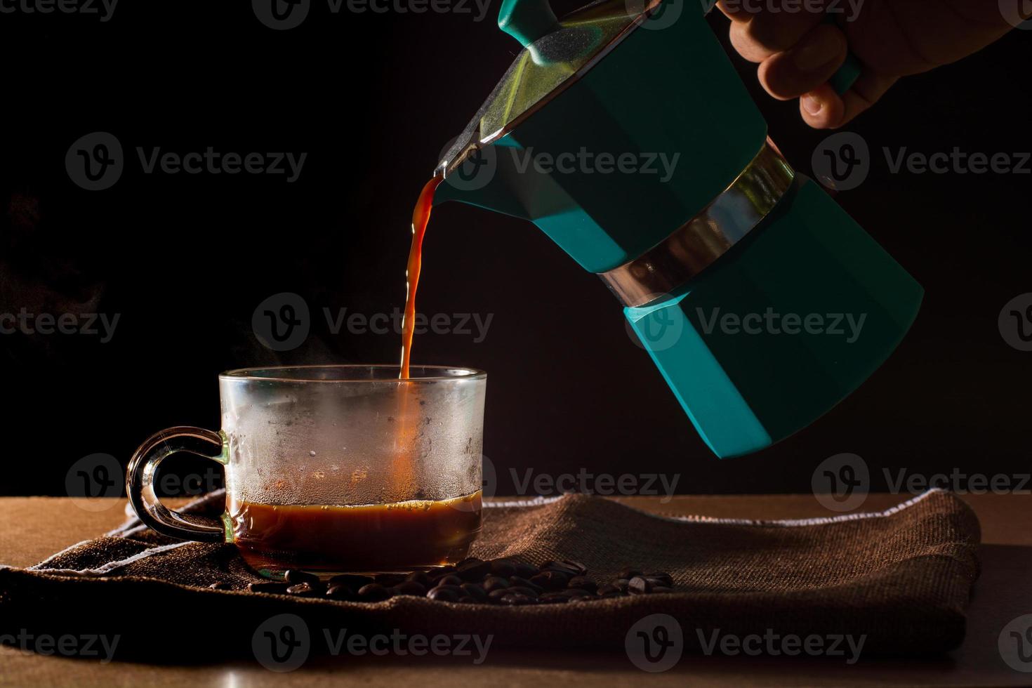 versez du café noir chaud du pot de moka vert pour effacer la tasse de café avec de la fumée et des grains de café sur une nappe marron et une table en bois. avantage du concept de café. photo